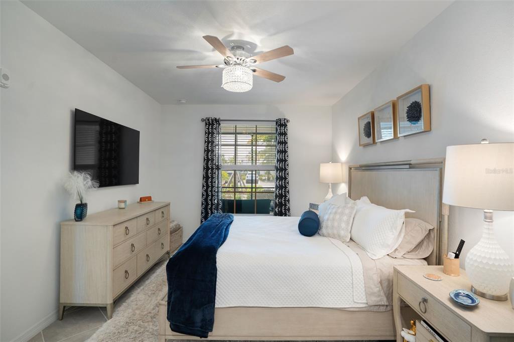 A bedroom features a bed, dresser, wall-mounted television, ceiling fan, and window with patterned curtains — 12440 WELLEN GOLF STREET UNIT 105, VENICE, FL