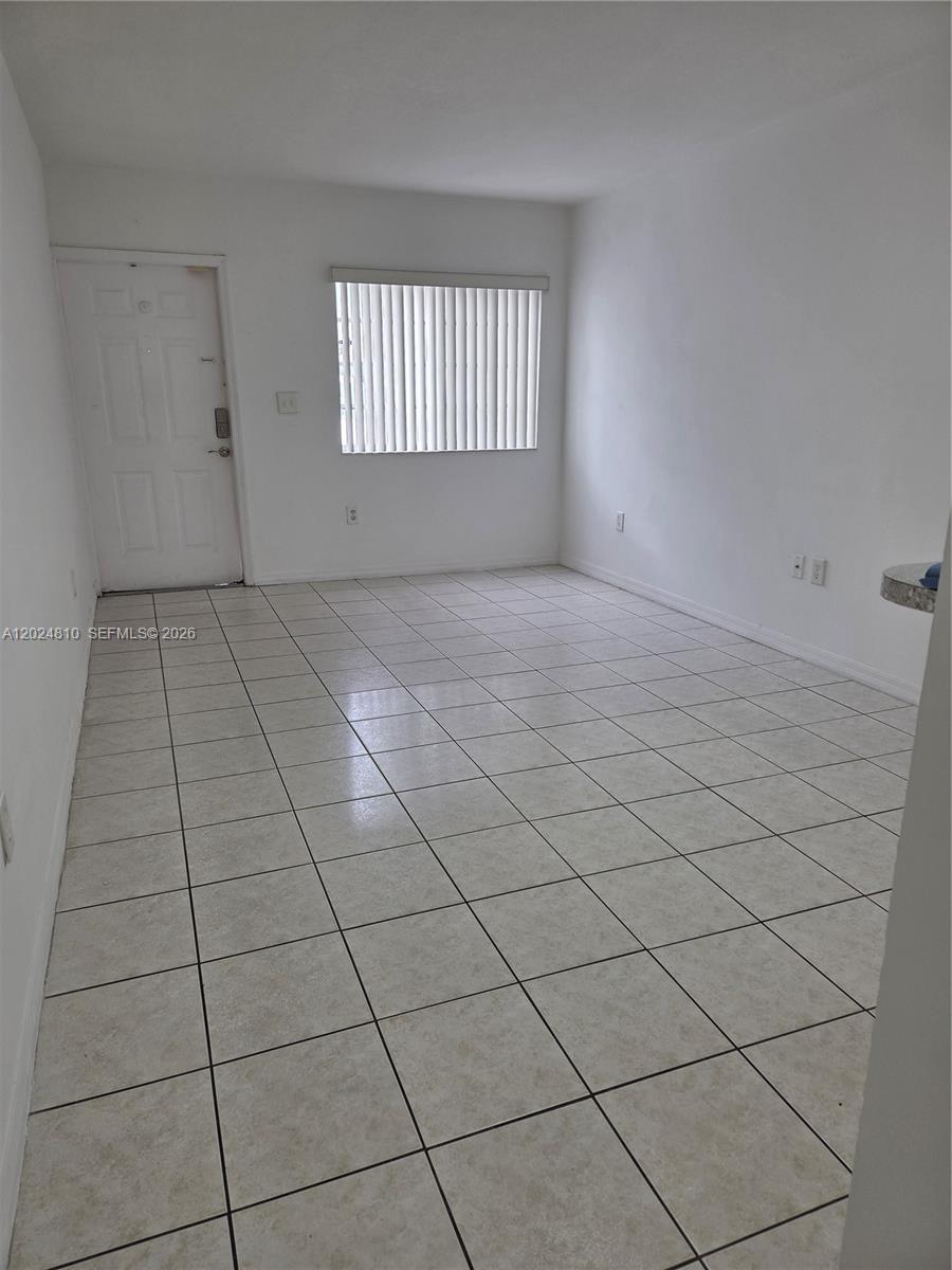 An empty room features white walls, light-colored square floor tiles, and a window with blinds — MIAMI, FL