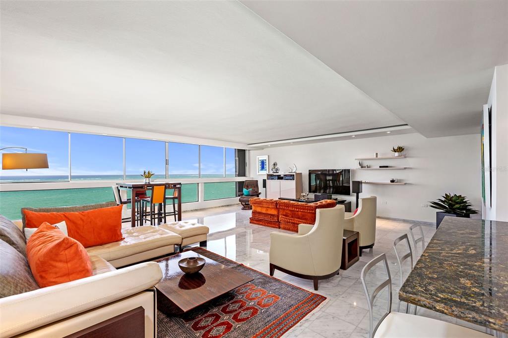 A modern living room features marble floors, orange furniture, and floor-to-ceiling windows overlooking the ocean — Plaza Atlantico AVE ISLA VERDE UNIT 1002, CAROLINA, PR