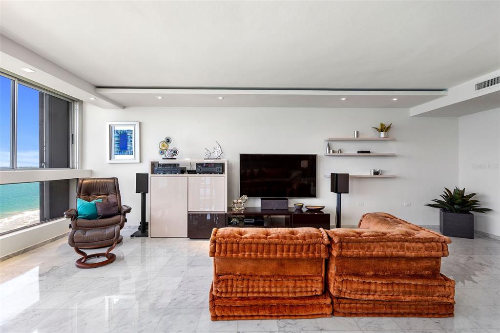 A living room features a large orange sectional, a television, and floor-to-ceiling windows overlooking water — Plaza Atlantico AVE ISLA VERDE UNIT 1002, CAROLINA, PR
