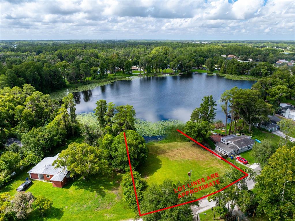 An aerial view shows a vacant grassy lot bordering a lake with surrounding residential homes — 21414 LAKE SHARON DRIVE, LAND O LAKES, FL