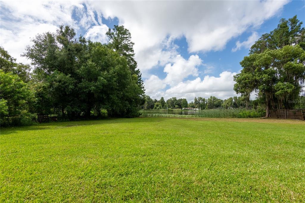 A large, grassy backyard leads to a lake surrounded by trees under a cloudy sky — 21414 LAKE SHARON DRIVE, LAND O LAKES, FL