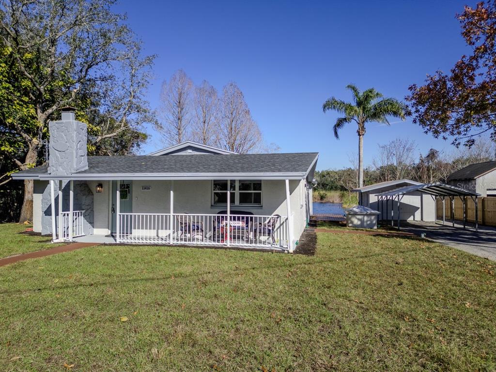 A single-story house with a stone chimney, front porch, and detached carport near a lake — 11416 PINE FOREST DRIVE, NEW PORT RICHEY, FL