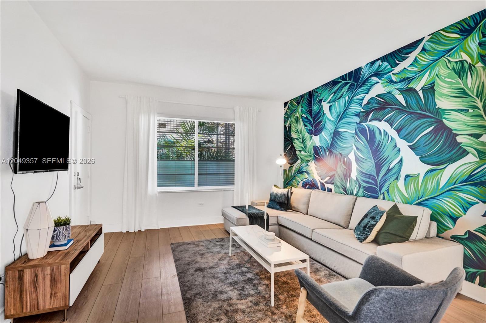 A living room features a large tropical leaf mural, a sectional sofa, and wood flooring — 1431 Lincoln Ter # 1, Miami Beach FL 33139, MIAMI BEACH, FL