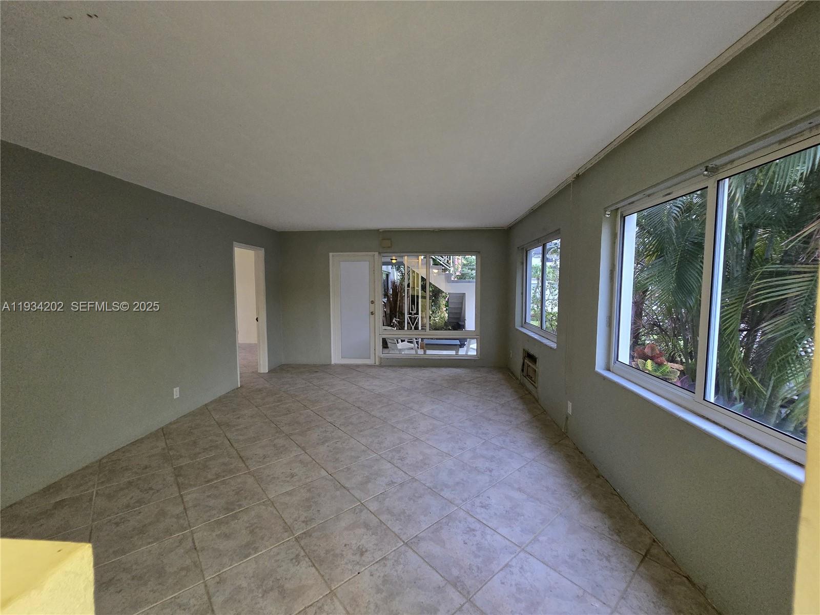 An empty room features light-colored tile flooring, gray walls, and large windows overlooking greenery — 3201 SE 12th St # B3, Pompano Beach FL 33062, POMPANO BEACH, FL