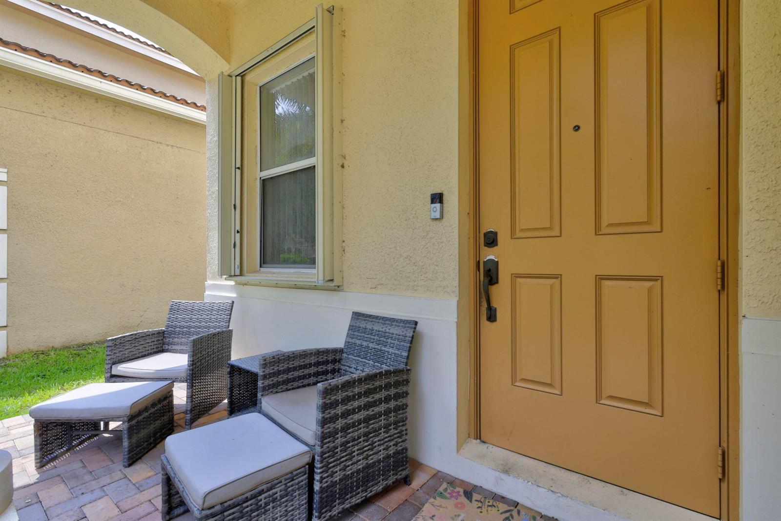 A patio with two wicker chairs and ottomans sits beside a yellow front door — 241 Gazetta Way, West Palm Beach FL 33413, WEST PALM BEACH, FL