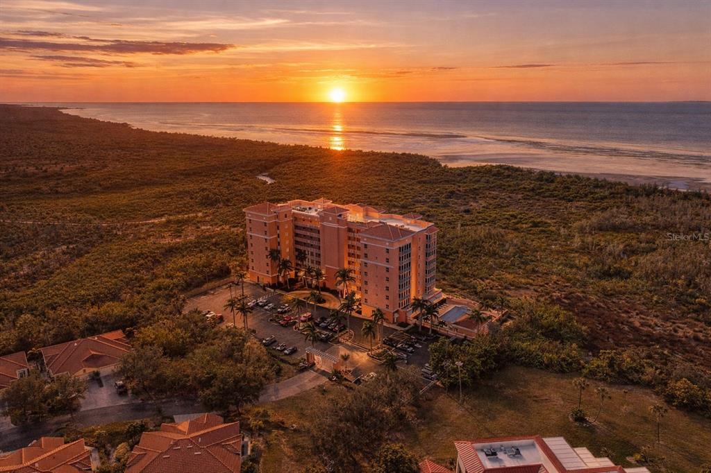 An aerial view shows a multi-story residential building surrounded by vegetation near a sunset beach — 3191 MATECUMBE KEY ROAD UNIT 307, PUNTA GORDA, FL