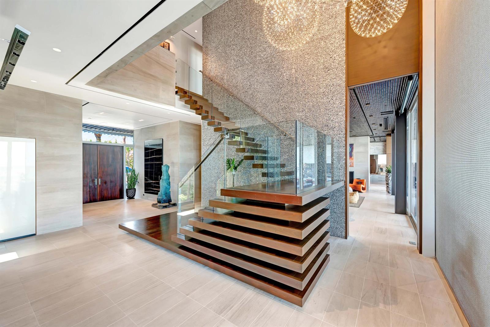 A modern foyer features a floating wooden staircase, glass railings, and a textured stone wall — VERO BEACH, FL