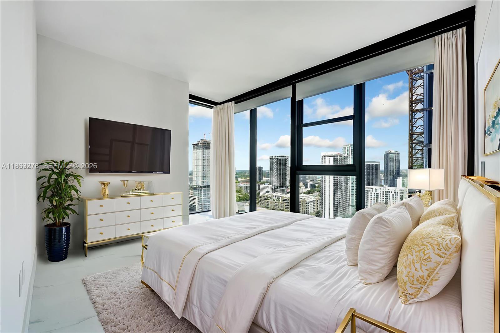 A modern bedroom features a white bed, a gold-accented dresser, and floor-to-ceiling windows overlooking city buildings — 725 NE 24th, Miami FL 33137, MIAMI, FL