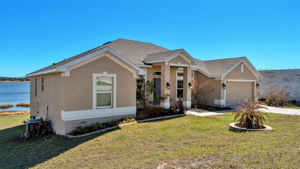 A tan, single-story suburban house with a two-car garage sits on a grassy waterfront lot — 4724 BROOKSHIRE COURT, LAKE WALES, FL