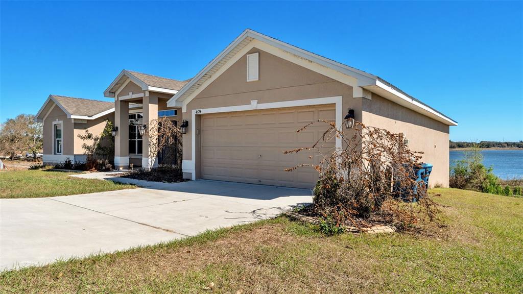 A tan single-story house with a two-car garage sits near a lake under blue skies — 4724 BROOKSHIRE COURT, LAKE WALES, FL