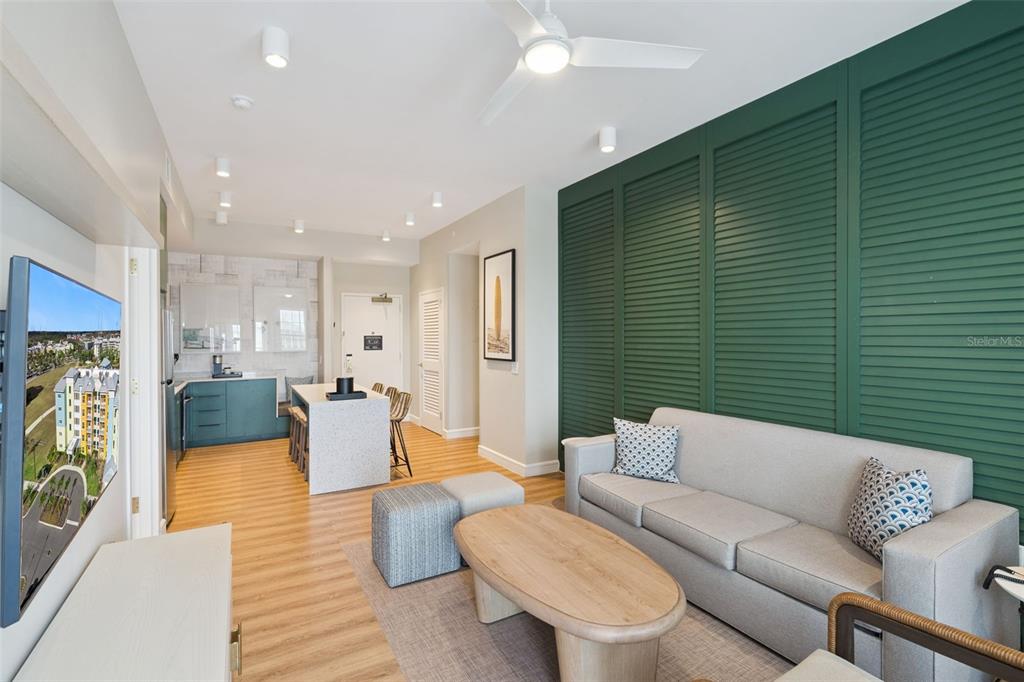 A modern living room and kitchen area features a green louvered wall and light wood flooring — 8061 VACATION AVENUE UNIT 2508, KISSIMMEE, FL