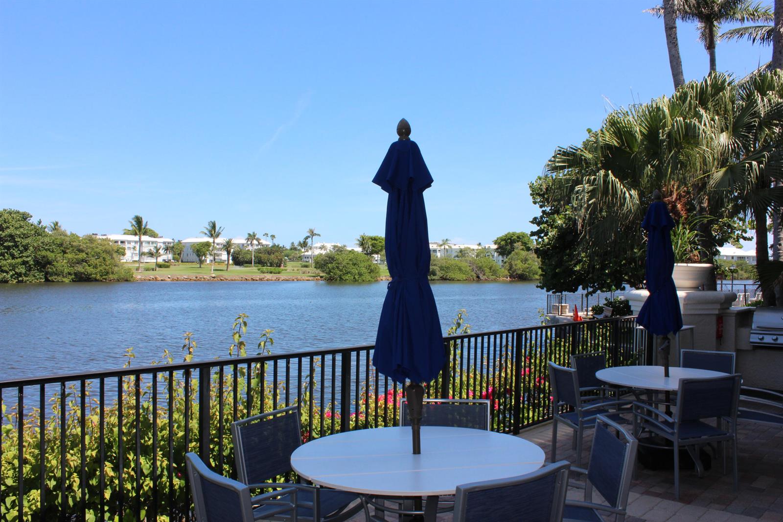 A patio with tables and blue umbrellas overlooks a lake surrounded by trees and buildings — 3302 Tuscany Way, Boynton Beach FL 33435, BOYNTON BEACH, FL