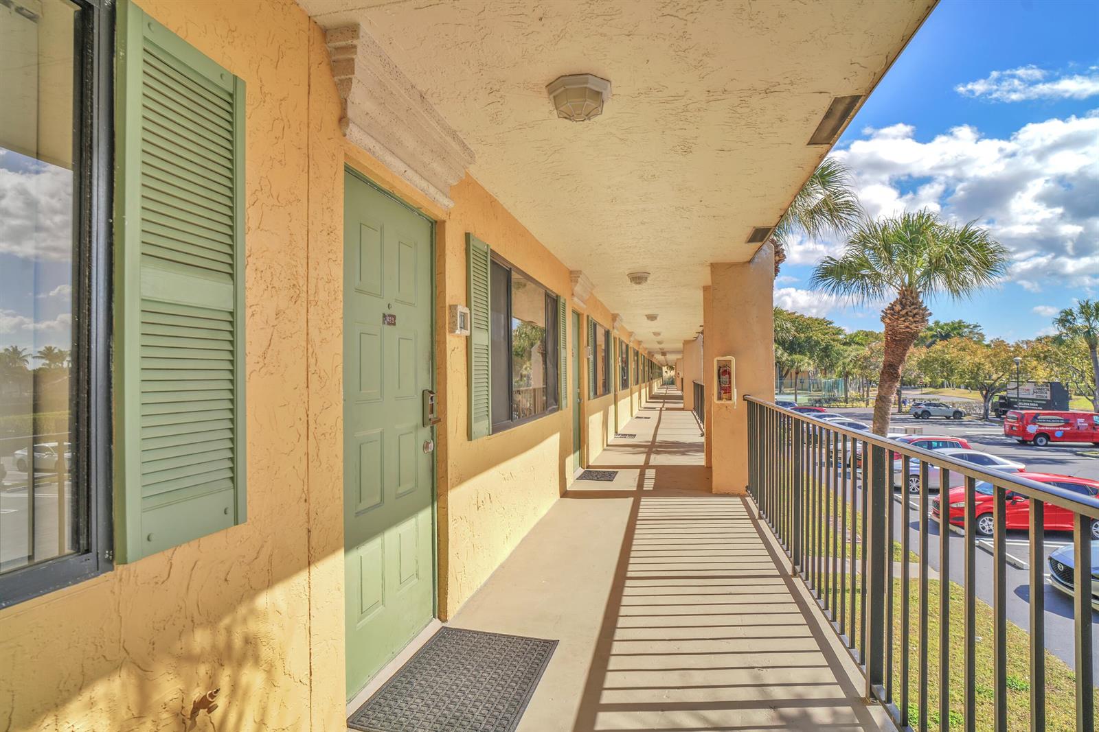 An outdoor walkway with green doors and shutters overlooks a parking lot with palm trees — 1423 Meadows Circle W, Boynton Beach FL 33436, BOYNTON BEACH, FL
