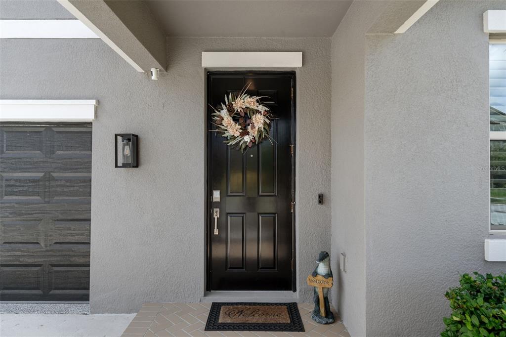 A black front door with a wreath and a welcome mat sits under a covered porch — 8755 FLOURISH DRIVE, LAND O LAKES, FL