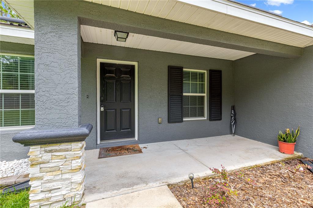 A covered front porch with a black door, stone-accented column, and a potted plant — 8707 SE 161ST STREET, SUMMERFIELD, FL