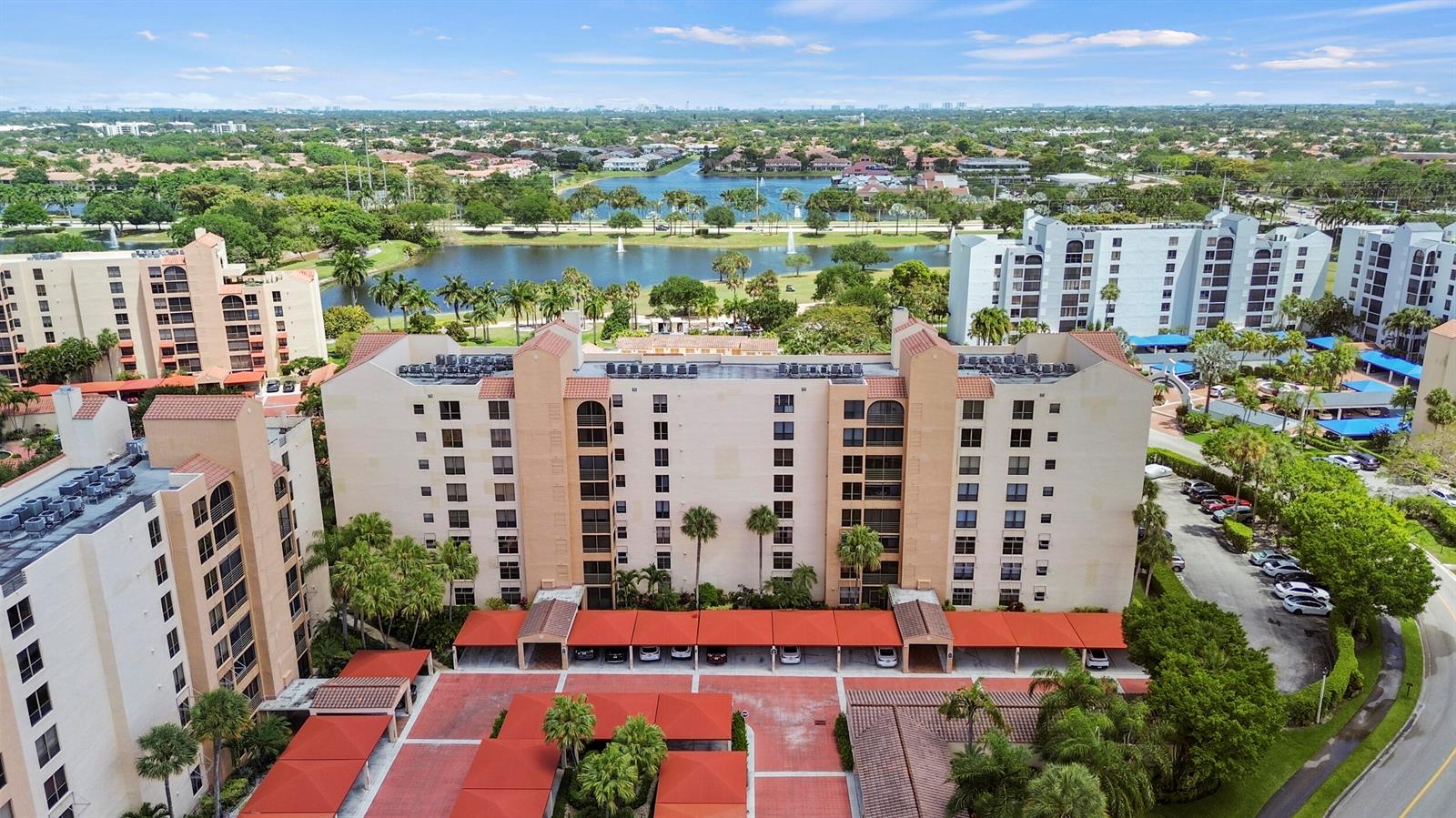 An aerial view shows multi-story apartment buildings with covered parking lots and a nearby lake — 7209 Promenade Drive # 101, Boca Raton FL 33433, BOCA RATON, FL