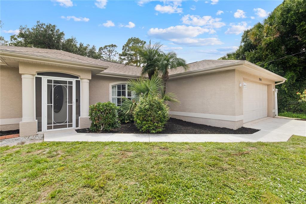 A beige single-story house features a white garage door, a screened entryway, and landscaping — 3941 SANTA ANA ROAD, NORTH PORT, FL