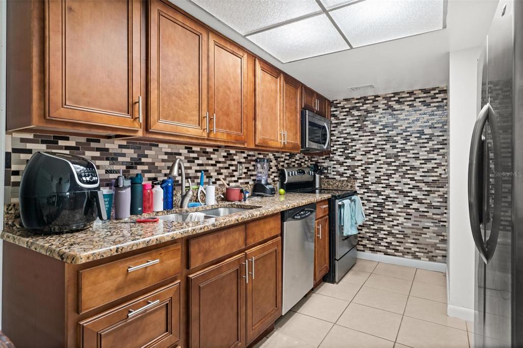 A kitchen features wood cabinets, granite countertops, a mosaic tile backsplash, and stainless steel appliances — 5201 VINELAND ROAD UNIT 112, ORLANDO, FL