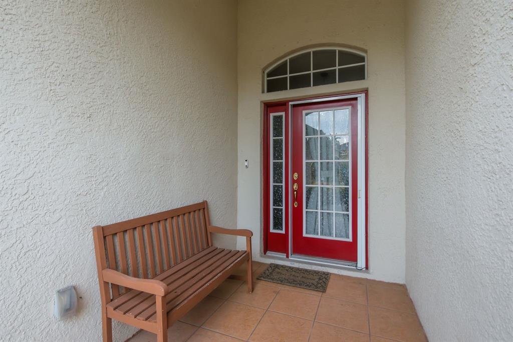 A wooden bench sits on a tiled porch in front of a red front door — 4882 W BREEZE CIRCLE, PALM HARBOR, FL