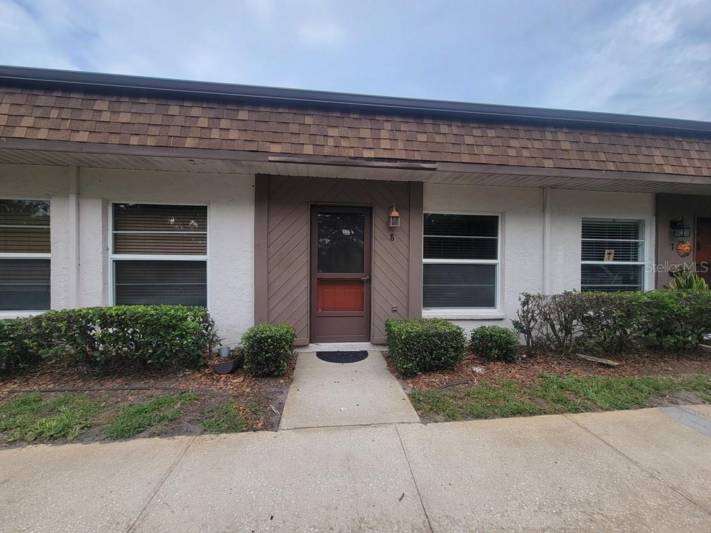 The exterior of a single-story residential unit features a brown door, shingled roof, and landscaping — 6141 CHESHAM DRIVE UNIT 8, NEW PORT RICHEY, FL