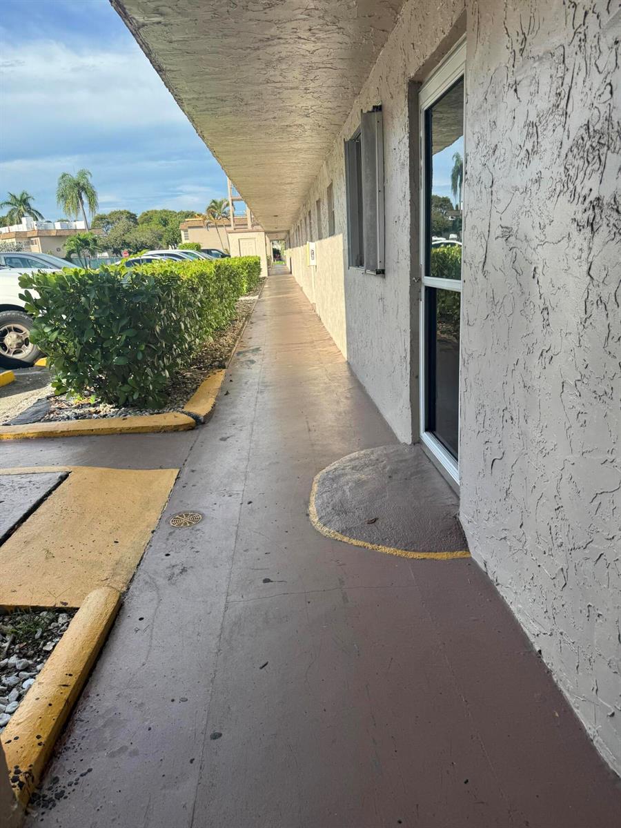 A concrete walkway runs alongside a building exterior with a glass door and landscaping — 725 Lori Drive # 107, Palm Springs FL 33461, PALM SPRINGS, FL
