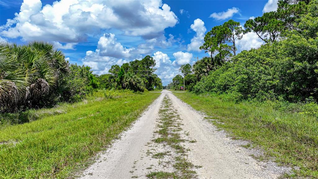 A dirt road stretches through a grassy, wooded landscape under a bright, cloudy blue sky — 1245 CAVALIER CIRCLE, LEHIGH ACRES, FL