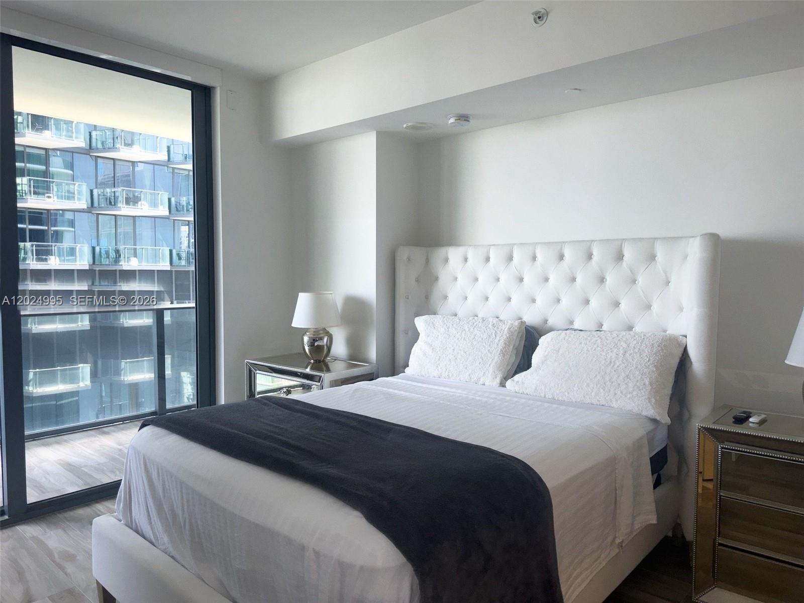 A bedroom features a tufted white headboard, mirrored nightstands, and a balcony with city views — 801 S Miami Ave # 2607, Miami FL 33130, MIAMI, FL