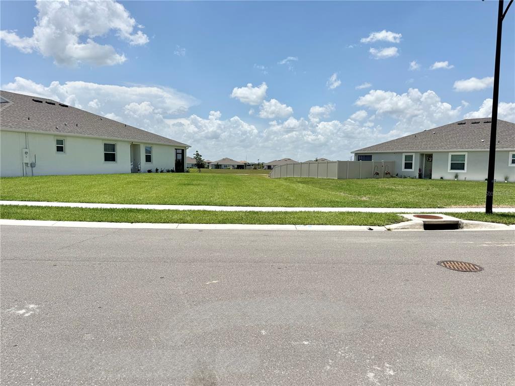 A vacant grassy lot sits between two suburban houses under a blue sky — 32828 RUSTIC RISE ROAD, SAN ANTONIO, FL