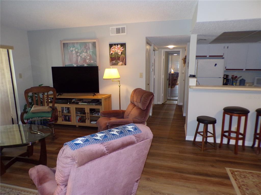A living room with wood-look flooring, a television console, two armchairs, and an adjacent kitchen — 19505 QUESADA AVENUE UNIT C207, PORT CHARLOTTE, FL