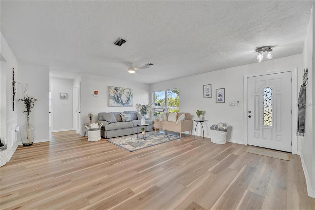 A bright living room features light wood-look flooring, a gray sofa, and a white front door — 6209 FLAMINGO DRIVE, APOLLO BEACH, FL
