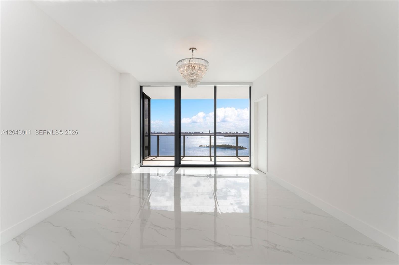 An empty room with white marble floors, a crystal chandelier, and floor-to-ceiling windows overlooking water — 700 NE 24 ST # 1004, Miami FL 33137, MIAMI, FL
