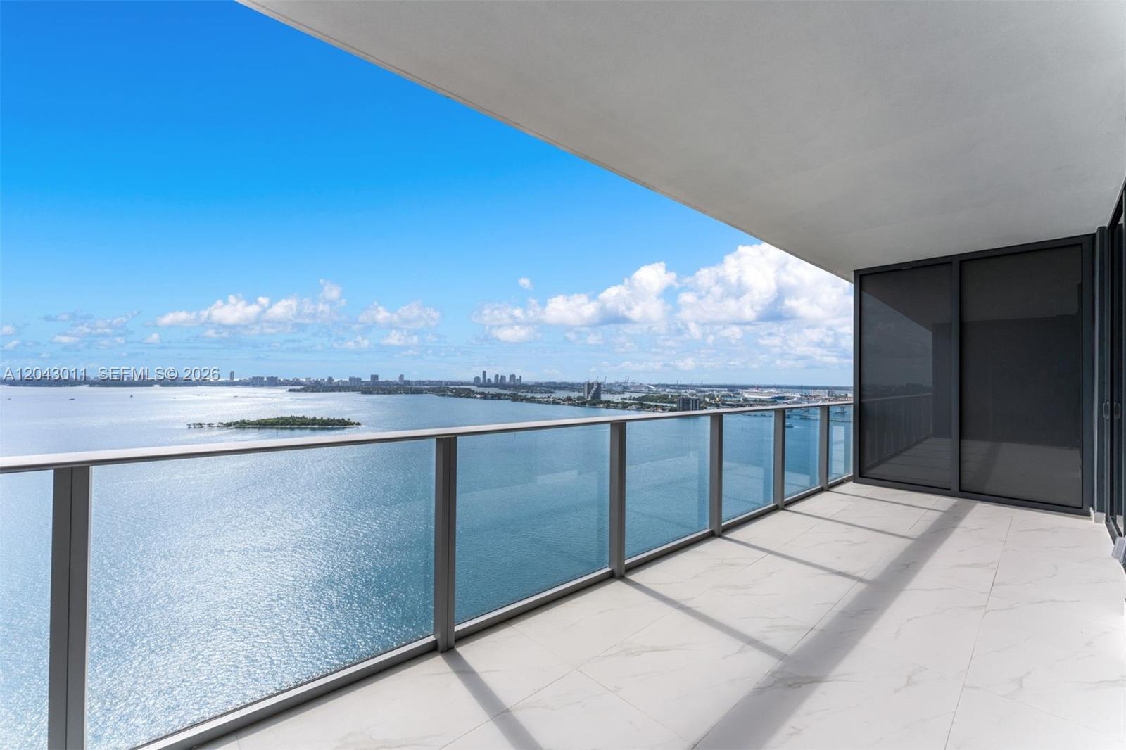 A modern balcony features white marble-look tile flooring and a glass railing overlooking the ocean — 700 NE 24 ST # 1004, Miami FL 33137, MIAMI, FL