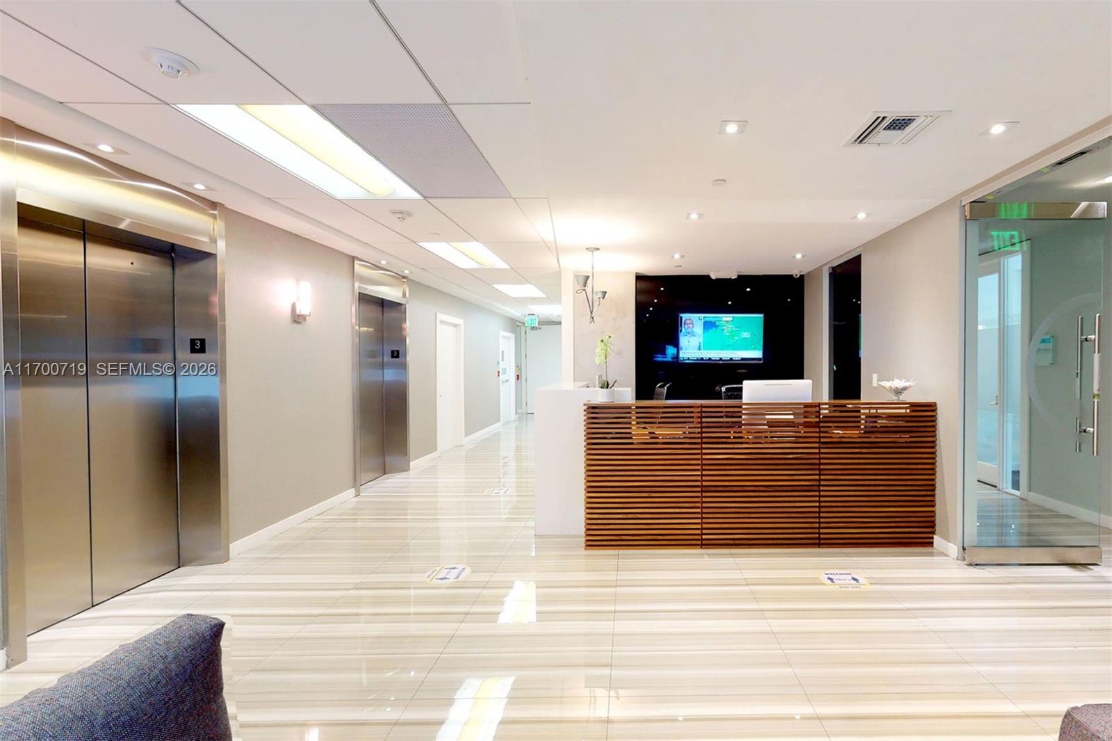 A modern office lobby features a wooden reception desk, stainless steel elevators, and tiled floors — 150 SE 2nd Ave # 319, FL 33131, FL