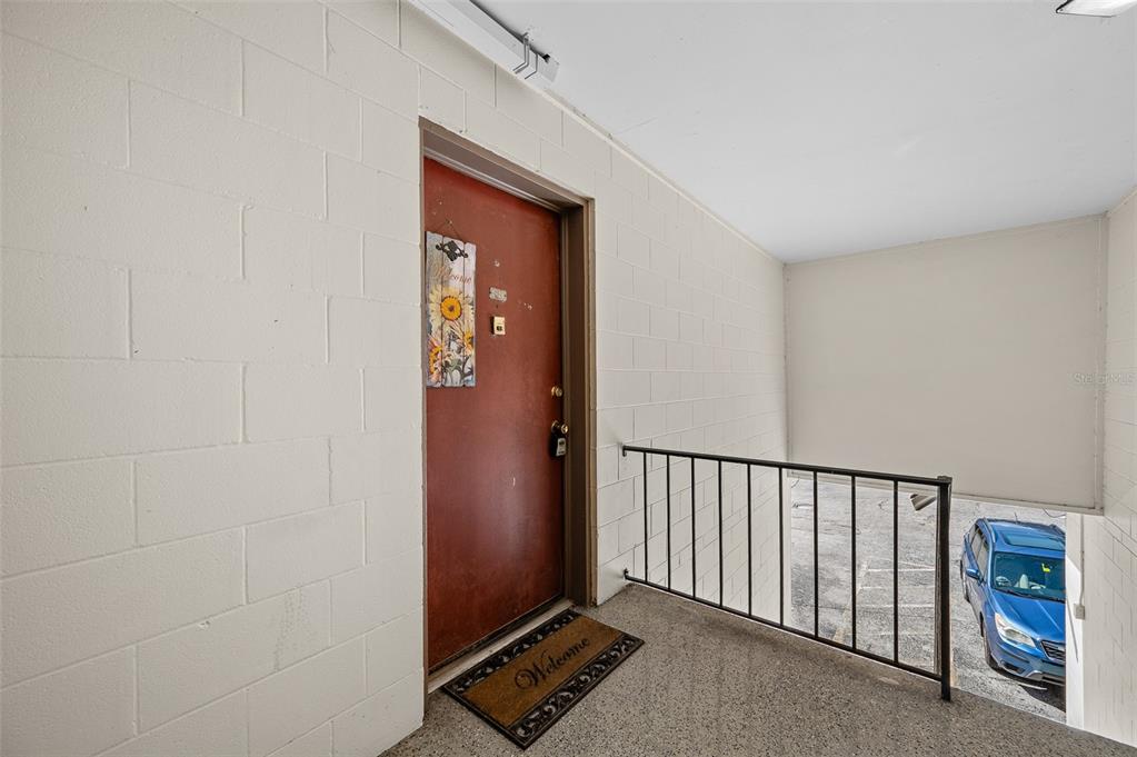 A red front door with a sunflower sign opens onto a covered concrete balcony — 1700 SW 16TH COURT UNIT D24, GAINESVILLE, FL