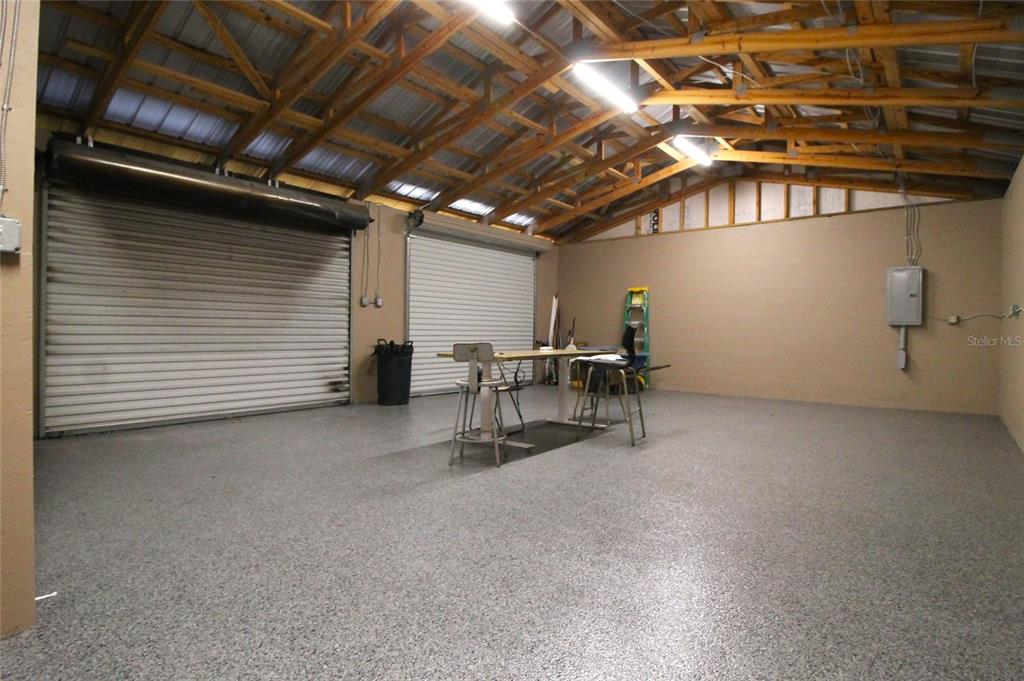 A spacious garage with two roll-up doors, exposed wooden trusses, and a speckled epoxy floor — 114 W MYERS BOULEVARD, MASCOTTE, FL