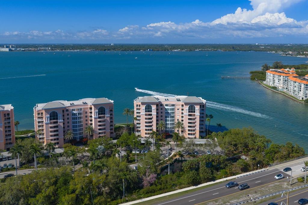 An aerial view shows two peach-colored waterfront condominium buildings near a road and bay — 4993 BACOPA LANE S UNIT 505, ST PETERSBURG, FL