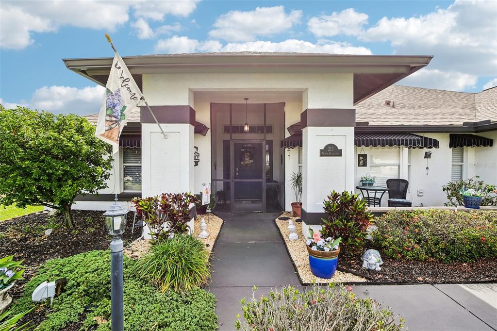 The exterior of a house features a covered entryway, a walkway, and landscaped garden beds — 213 N BROCKFIELD DRIVE, SUN CITY CENTER, FL