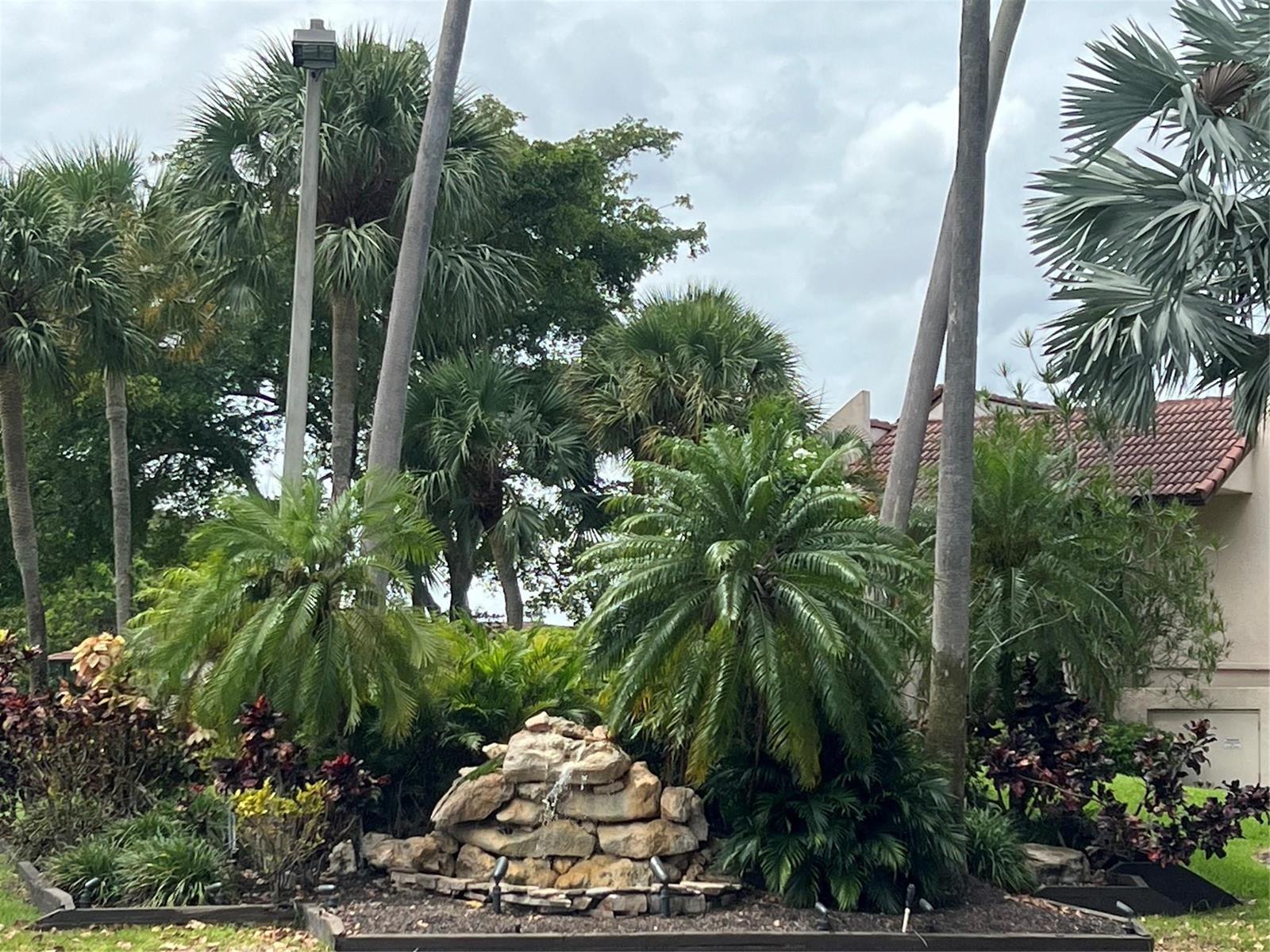A rock water feature sits in a landscaped garden with palm trees and a house — 21542 Juego Circle # 27h, Boca Raton FL 33433, BOCA RATON, FL