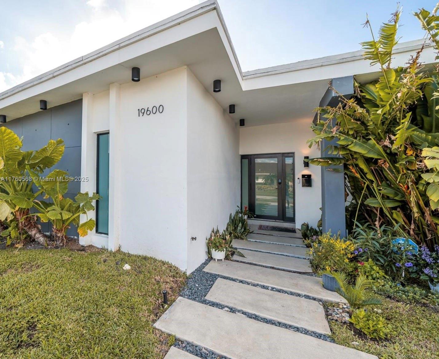 A modern house exterior features a white facade, a concrete walkway, and lush tropical landscaping — 19600 NE 19th Ave, Miami FL 33179, MIAMI, FL