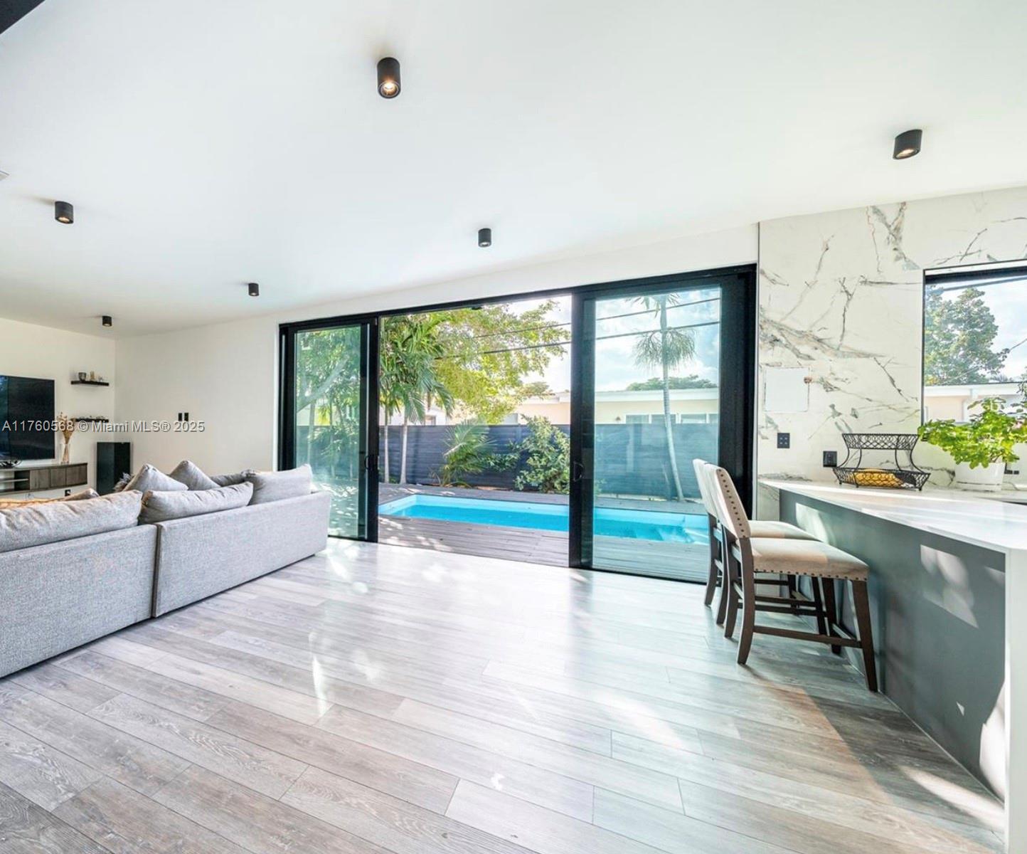 A modern living room and kitchen area features sliding glass doors opening to a backyard pool — 19600 NE 19th Ave, Miami FL 33179, MIAMI, FL