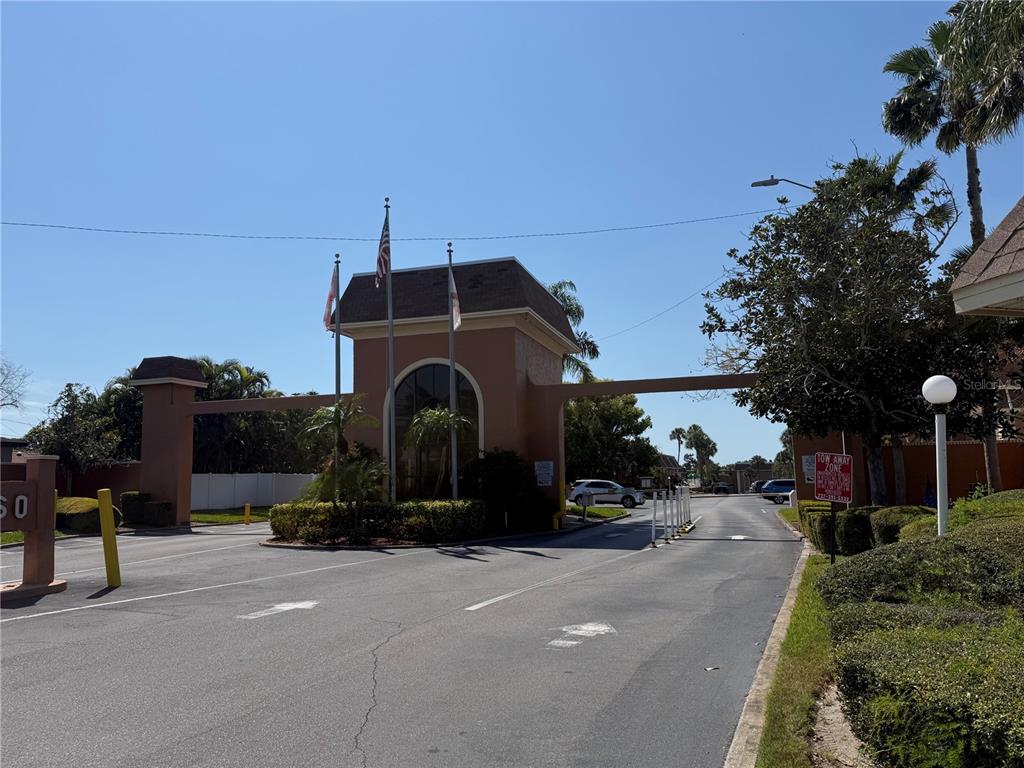 A gated entrance with a tan structure, flags, and a paved road under a clear sky — 12760 INDIAN ROCKS ROAD UNIT 1024, LARGO, FL