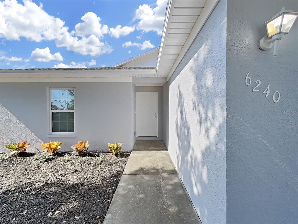 A concrete walkway leads to the white front door of a light gray stucco house — 6240 ARTHUR AVENUE, NEW PORT RICHEY, FL
