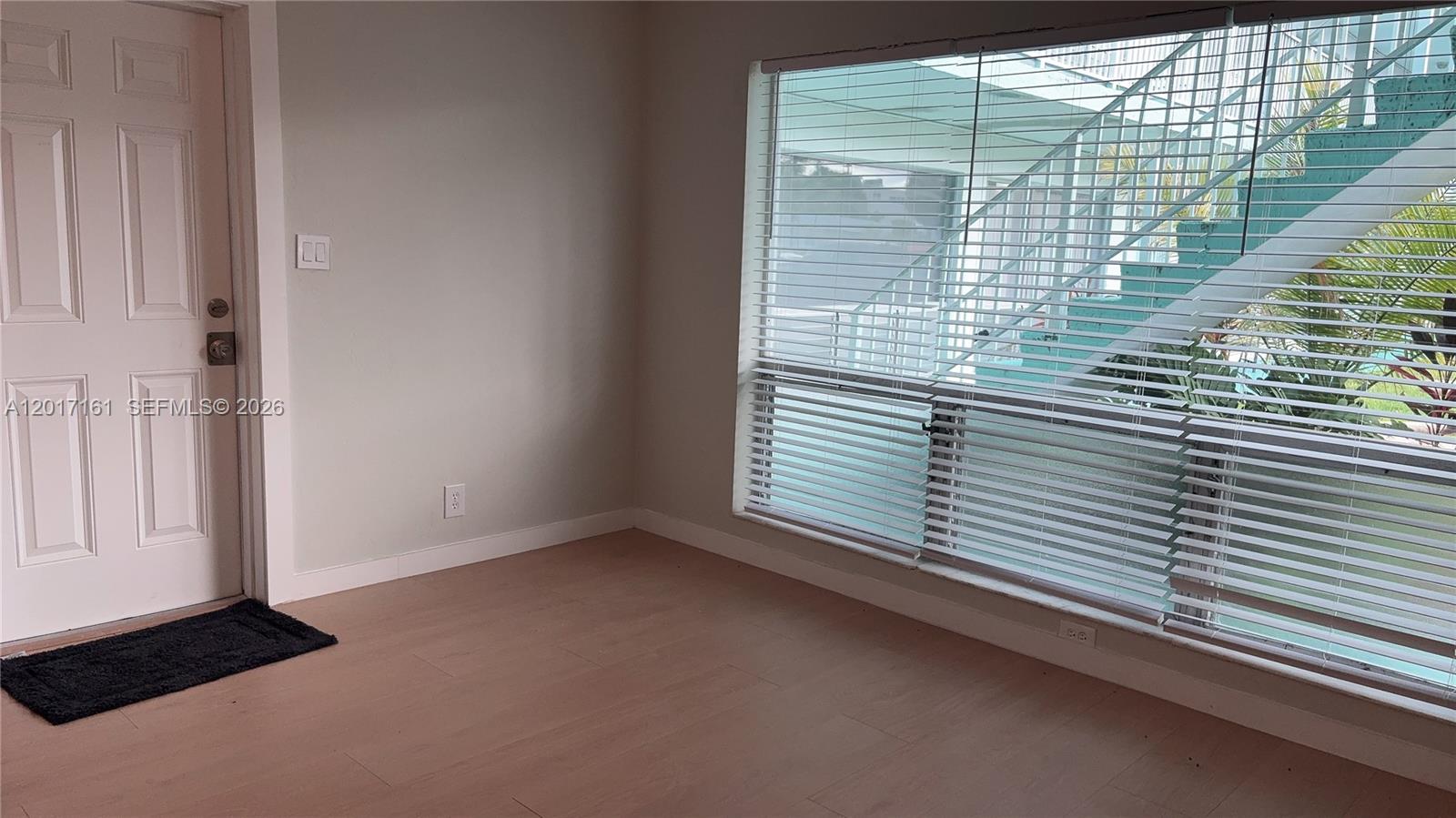 An empty room features light wood-look flooring, a white door, and a large window with blinds — 3212 NE 7th Pl # 10, Pompano Beach FL 33062, POMPANO BEACH, FL