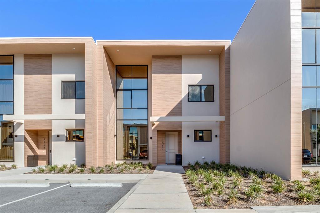 A modern two-story townhouse exterior features a tall glass window, light-colored siding, and concrete walkway — 7540 BROOKLYN DRIVE, KISSIMMEE, FL