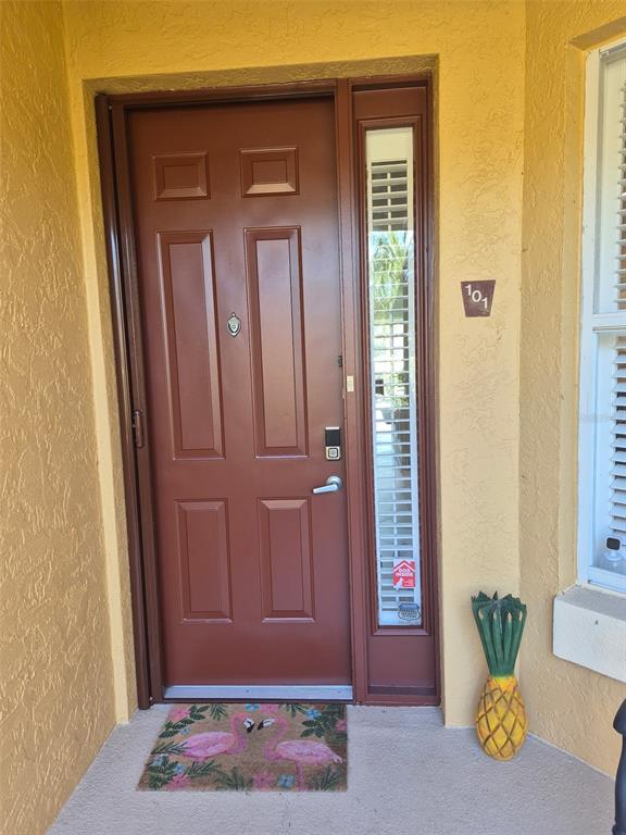 A brown front door with a side window and a flamingo doormat at the entrance — 7179 BOCA GROVE PLACE UNIT 101, BRADENTON, FL