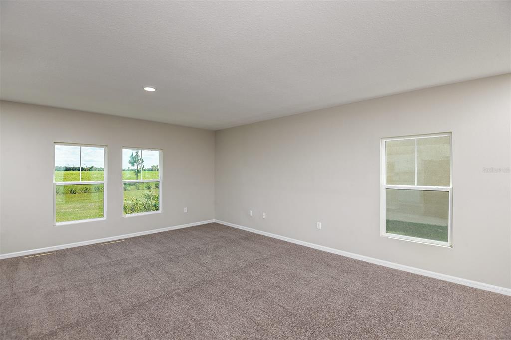 An empty room with beige walls, gray carpeting, and three windows overlooking a grassy field — 35433 ARBOR RETREAT DRIVE, DADE CITY, FL