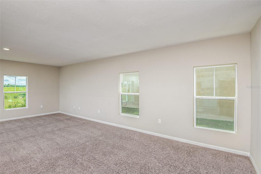 An empty room with beige walls, gray carpeting, and three windows overlooking a grassy area — 35433 ARBOR RETREAT DRIVE, DADE CITY, FL