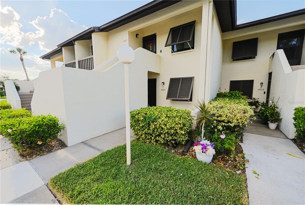 A two-story apartment building exterior features light-colored stucco walls, dark shutters, and a front walkway — 4625 LONGWATER CHASE UNIT 113, SARASOTA, FL