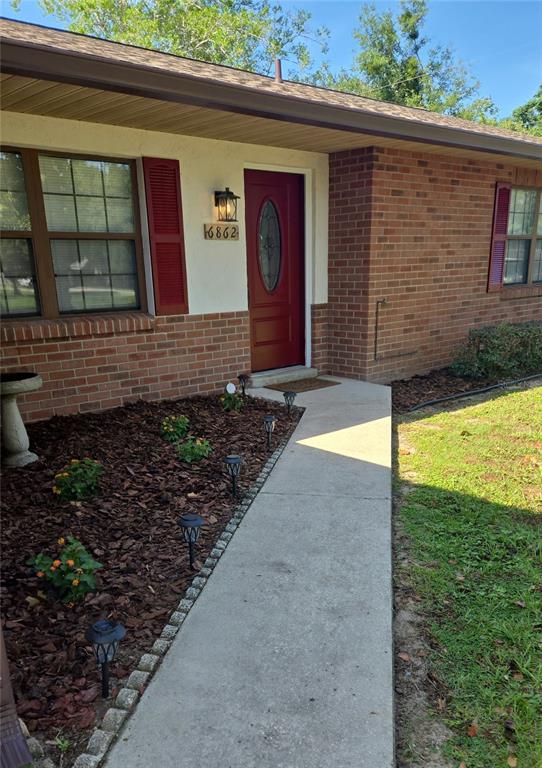 A brick house exterior features a red front door, a concrete walkway, and landscaped mulch beds — 6862 SE 52 PLACE, OCALA, FL